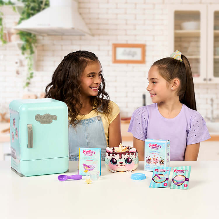 Two girls enjoying the Cookeez Makery S2 Freezy Cakez Playset with a colorful cake and play accessories.
