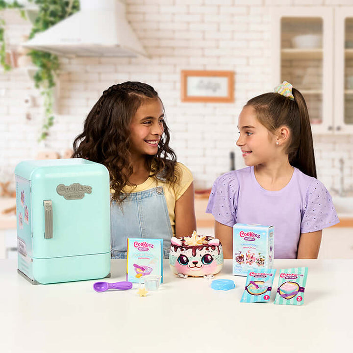 Two girls enjoying the Cookeez Makery S2 Freezy Cakez Playset with a colorful cake and play accessories.